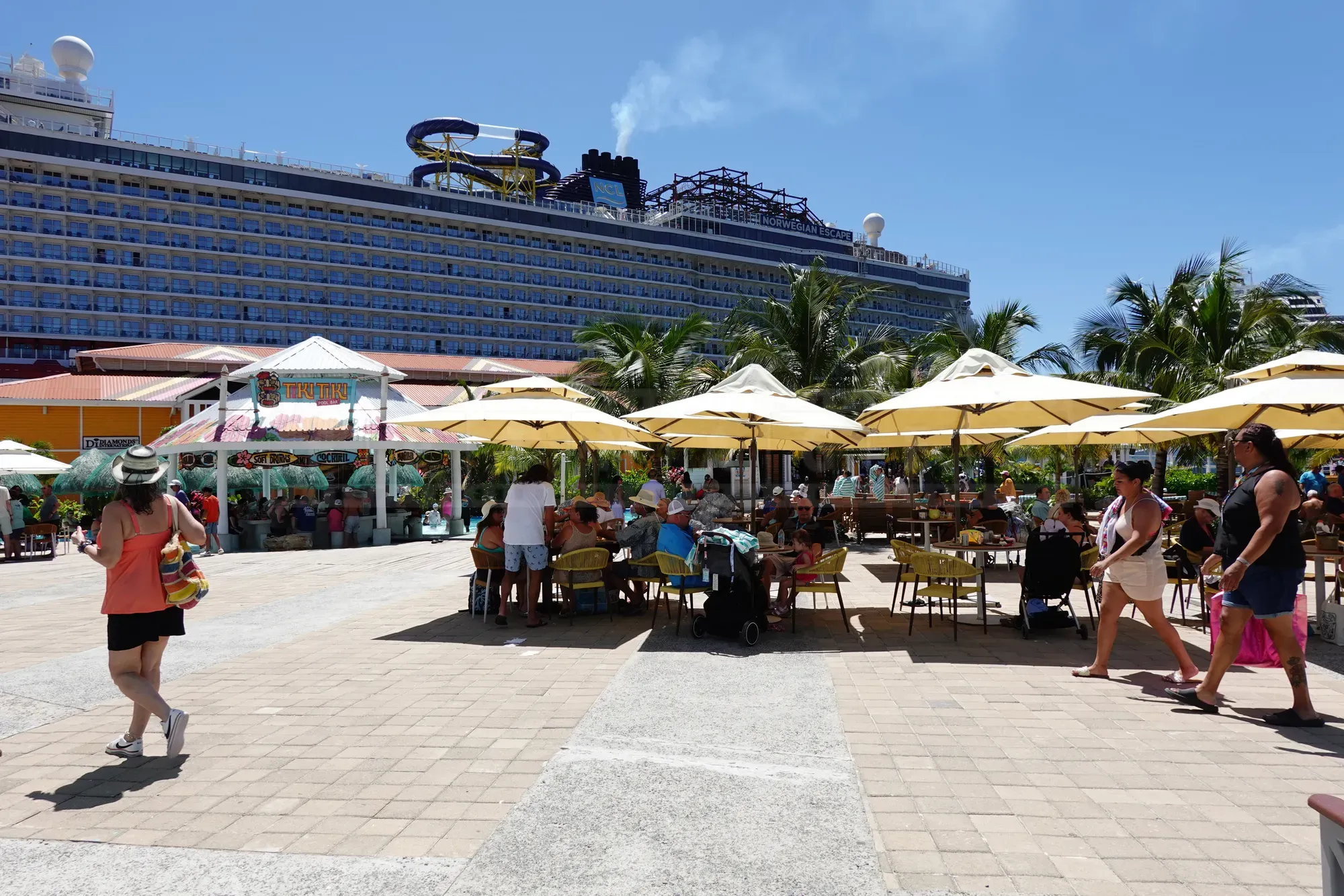 Outdoor seating area with yellow umbrellas at the Coxen Hole cruise port in Roatán, with the Norwegian Escape cruise ship docked behind palm trees.